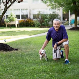 A man crouching on a grassy lawn with a small dog in an outdoor setting.