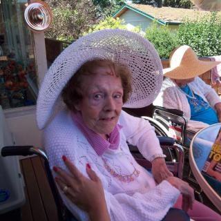 An elderly woman wearing a sun hat sits outdoors in a wheelchair.