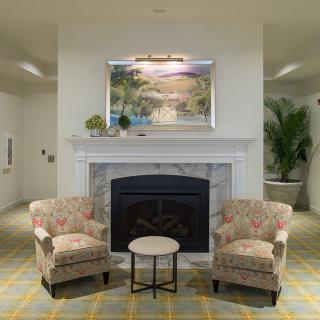 A cozy living room setup featuring a fireplace flanked by two floral-patterned chairs and a small round table, with artwork and greenery adding to the ambiance.