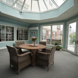 A sunlit conservatory dining area featuring a round table and wicker chairs under a glass roof.