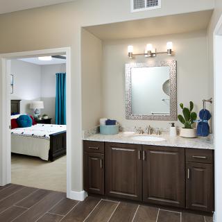 A modern bathroom with a vanity area featuring a mirror, sink, and dark wood cabinets. The adjacent bedroom is visible through an open doorway.