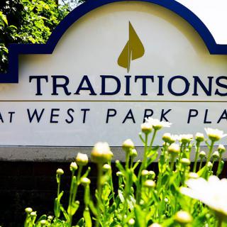 A sign for Traditions at West Park Place surrounded by greenery and flowers.