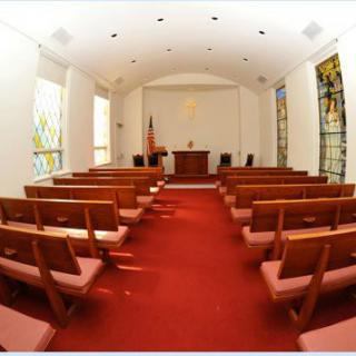 A small chapel interior with wooden pews, a red carpet, and stained glass windows.