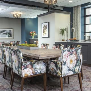 A modern dining room with a large wooden table surrounded by floral-patterned chairs, illuminated by chandeliers and framed by large windows.