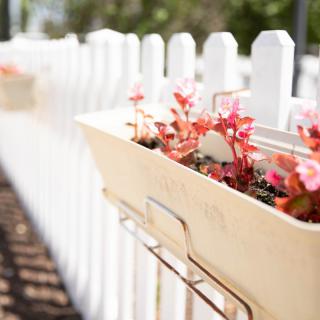 A planter filled with pink flowers hangs on a white picket fence.