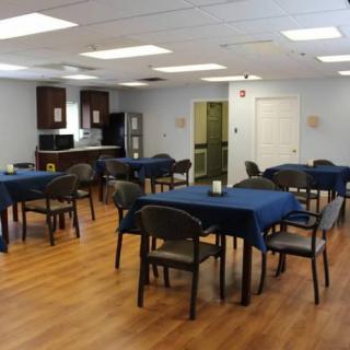 A dining area with multiple tables covered in blue tablecloths and chairs, featuring a kitchen setup and wooden flooring.