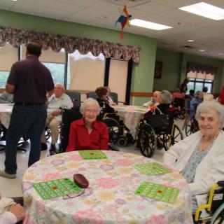 Elderly individuals gathering in a community room, some seated in wheelchairs and playing bingo.