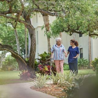 A caregiver walking with an elderly person in a garden setting.