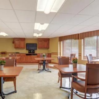 A well-lit dining area with wooden tables and chairs, featuring a television against one wall and large windows allowing natural light inside.