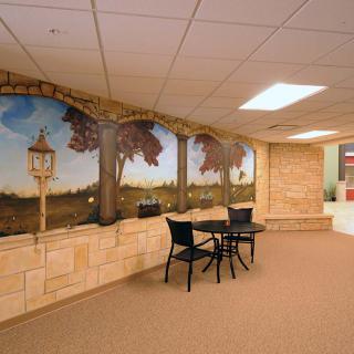 A cozy indoor seating area featuring a mural depicting an outdoor scene, with a small table and two chairs along a decorative brick wall.