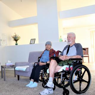 An elderly couple sits together in a living room, one in a wheelchair, enjoying a peaceful moment indoors.