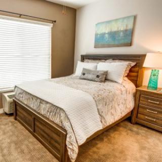 A cozy bedroom featuring a neatly made bed with decorative pillows, flanked by wooden nightstands and a lamp. A scenic painting hangs above the headboard, and natural light filters through the window blinds.