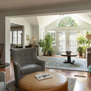 A cozy living room with comfortable armchairs, a round coffee table, and large windows allowing ample natural light. The space is decorated with plants and features a hardwood floor.