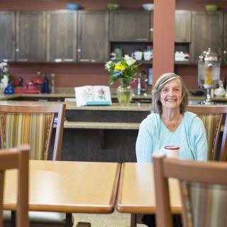 A woman sits smiling in a cozy cafe with a kitchen area in the background.