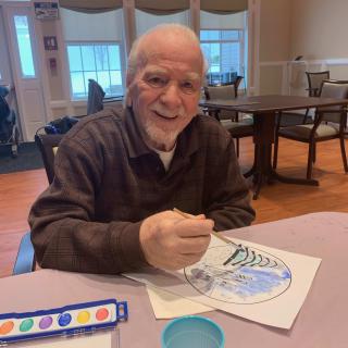 An elderly man smiling while engaging in a painting activity indoors.