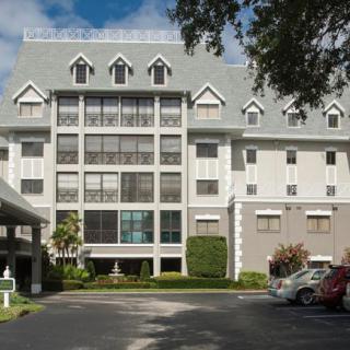 A multi-story building with a parking area and surrounding trees under a partly cloudy sky.