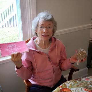 An elderly woman joyfully holds a card while sitting at a table.