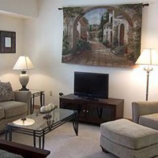 A cozy living room featuring a comfortable sofa set, a television on a stand, and ambient lighting from lamps. A decorative artwork hangs on the wall above the TV.
