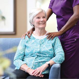 A senior woman sits comfortably while a healthcare professional provides support.