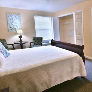 A cozy bedroom with a neatly made bed, two chairs, and a lamp by the window.