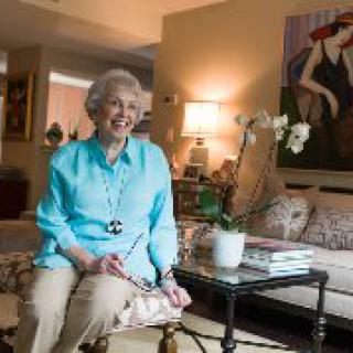 An elderly woman smiles while seated in a cozy living room, surrounded by artwork and furnishings.