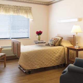 A clean and well-organized hospital room featuring a bed, bedside table with flowers, a lamp, and a chair next to a window.