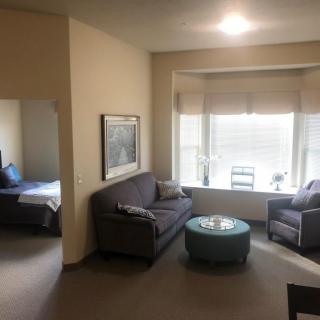 A cozy living room with a sofa, armchair, and a window, adjacent to a bedroom.