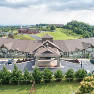 Aerial view of a large building surrounded by greenery and parking areas under a cloudy sky.