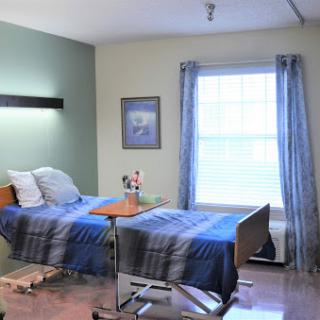 A well-lit patient room featuring two hospital beds, a window with blue curtains, and decorative artwork on the wall.