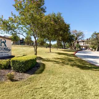 A landscaped area with a sign, trees, grass, and a road leading to a building.