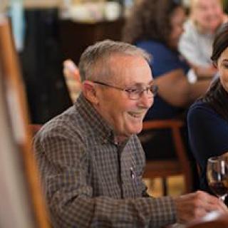 An elderly man with glasses enjoys a meal and laughs with others at a restaurant.