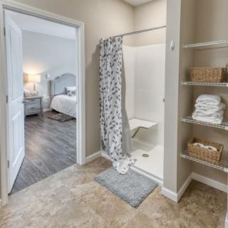 A modern bathroom with a shower and neatly organized shelves, leading into a cozy bedroom.
