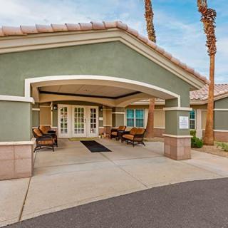 A welcoming building entrance featuring a covered patio with seating and palm trees in the background.