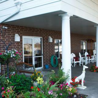 A cozy porch with white columns and a variety of colorful flowers, featuring a small statue and seating area.
