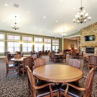 A spacious dining room with wooden tables and chairs, decorated with chandeliers and a carpeted floor, featuring large windows, a stone fireplace, and a television.