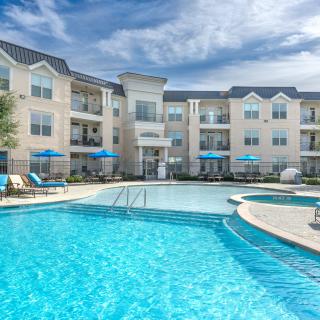 A modern apartment complex featuring a large outdoor swimming pool with lounge chairs under blue skies.