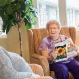 An elderly woman sitting comfortably in a chair, holding a magazine and smiling.