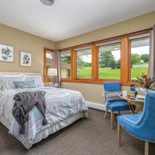 A cozy bedroom with a large window overlooking a green landscape, featuring a bed and blue chairs.