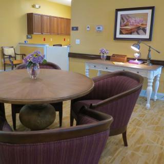 A cozy dining area with a round table, cushioned chairs, and a small desk with a lamp, set adjacent to a kitchen.