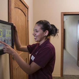A woman in a uniform interacts with a touchscreen device indoors, smiling as she works.