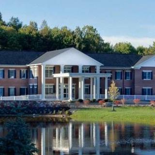A beautiful building with a white facade and large windows is reflected in a calm lake, surrounded by lush greenery.