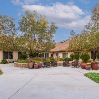 A peaceful courtyard with outdoor seating, surrounded by trees and potted plants under a partly cloudy sky.