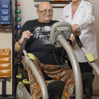 An elderly man exercising on a machine with the assistance of a healthcare professional.