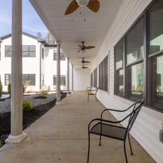 A long porch with chairs, ceiling fans, and large windows along a white building.