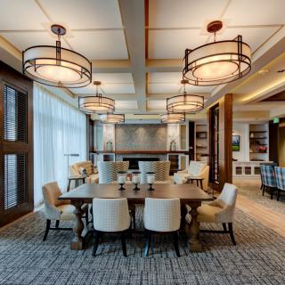 A modern dining room with a large wooden table, surrounded by upholstered chairs and elegant chandeliers overhead.
