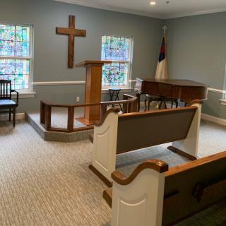 A small chapel interior featuring a pulpit, wooden pews, and stained glass windows.