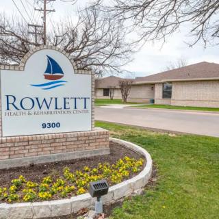 A sign for Rowlett Dental Associates stands in front of a clinic building, surrounded by flowers and a road.