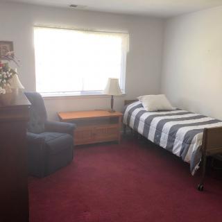 A simple bedroom with a single bed, chair, and window letting in natural light.