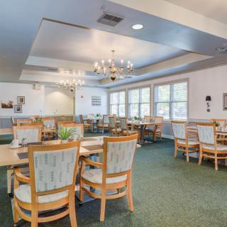 A well-lit dining room with multiple tables and chairs, featuring a chandelier and large windows.