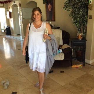A woman in a white dress stands in a living room holding some items.